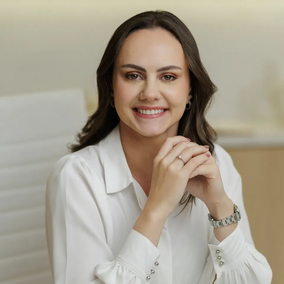 Dra. Larissa Basmage sentada à mesa, de blusa branca, com as mãos unidas e sorrindo